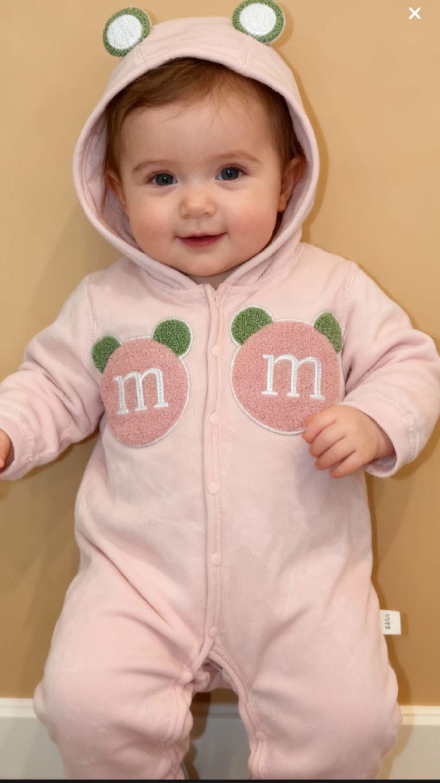 Baby wearing a pink onesie with M&M logos and frog eyes on a beige background