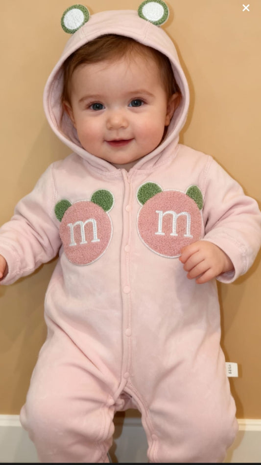 Baby wearing a pink onesie with M&M logos and frog eyes on a beige background