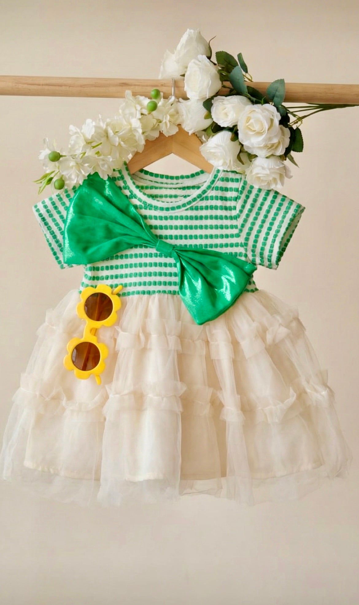 Green and white checkered dress with a large bow, white flowers, and yellow sunglasses on a wooden hanger.