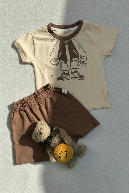 Children's outfit with beige shirt and brown shorts, featuring a teddy bear toy, on a light gray background.