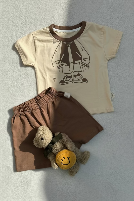 Children's outfit with beige shirt and brown shorts, featuring a teddy bear toy, on a light gray background.